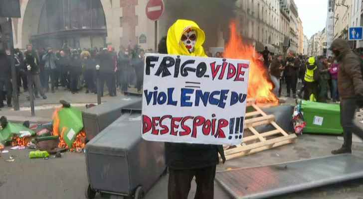 Masked demonstrator posing in front of burnt out bins with a sign reading "empty fridge = violence of desperation!"